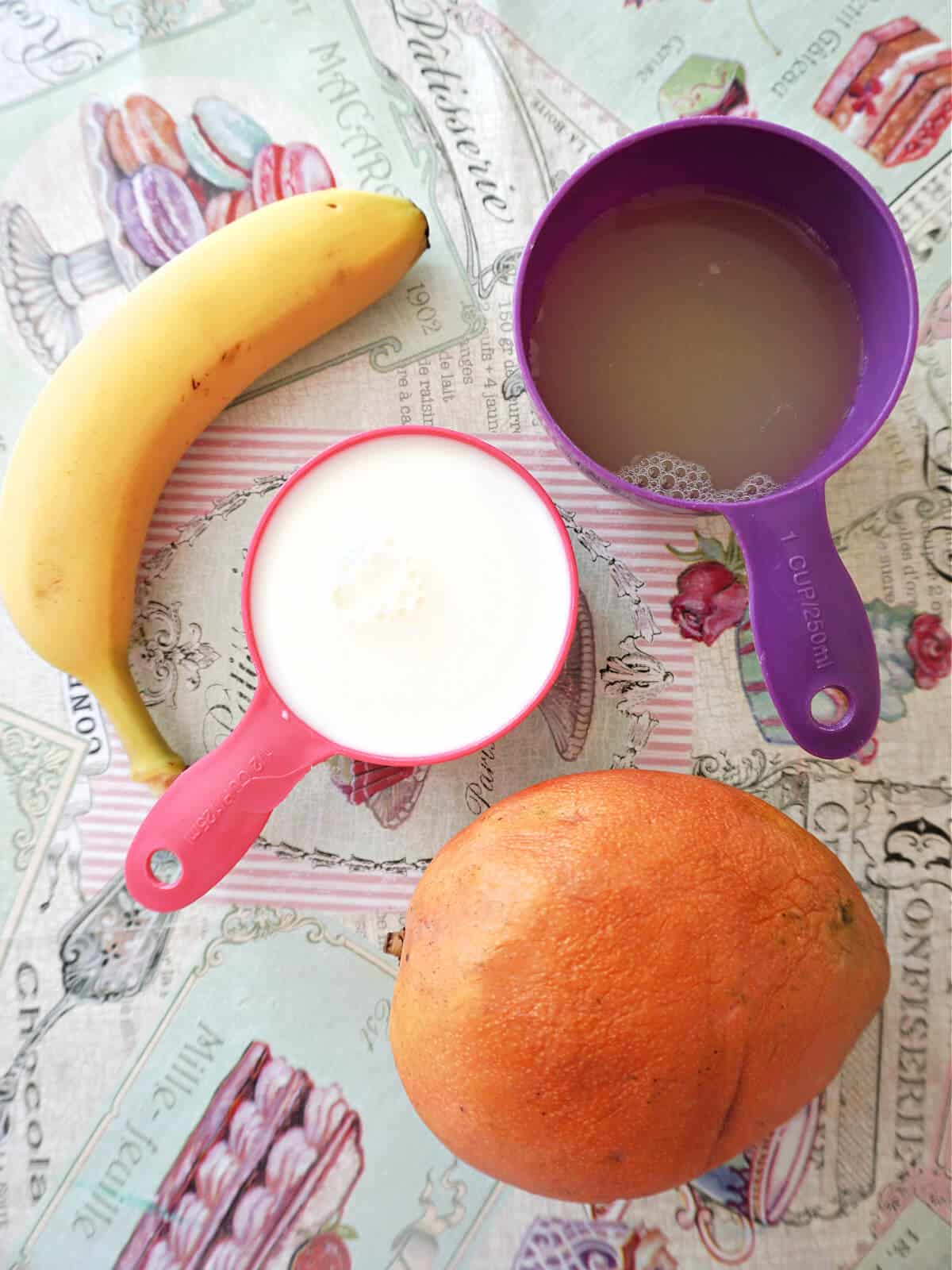 Overhead shoot of 1 mango, 1 banana, a cup of milk and a cup of juice.