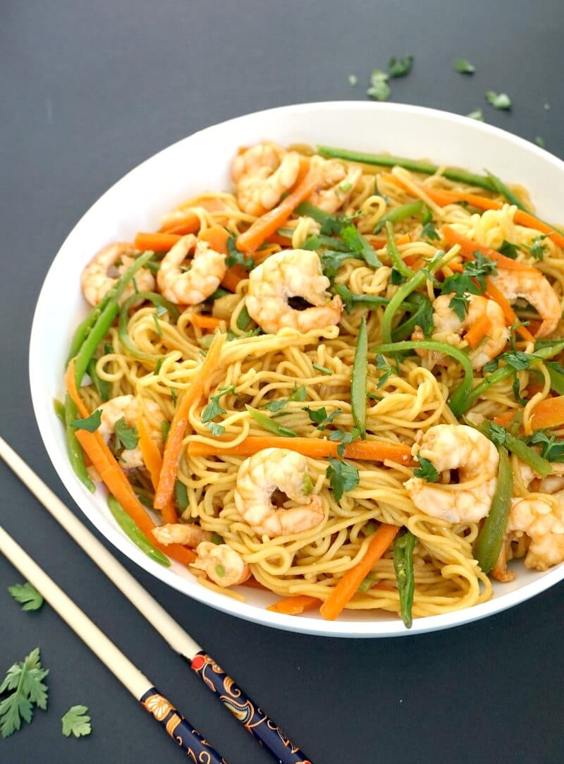 A white plate with shrimp stir fry and noodles and chop sticks on the side