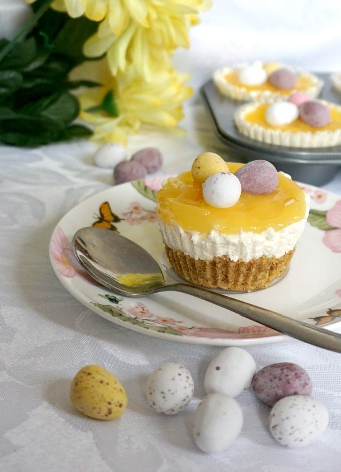 A mini lemon cheesecake on a small plate with a teaspoon next to it, easter eggs scattered around, and yellow flowers and a tray of mini lemon cheesecakes in the background