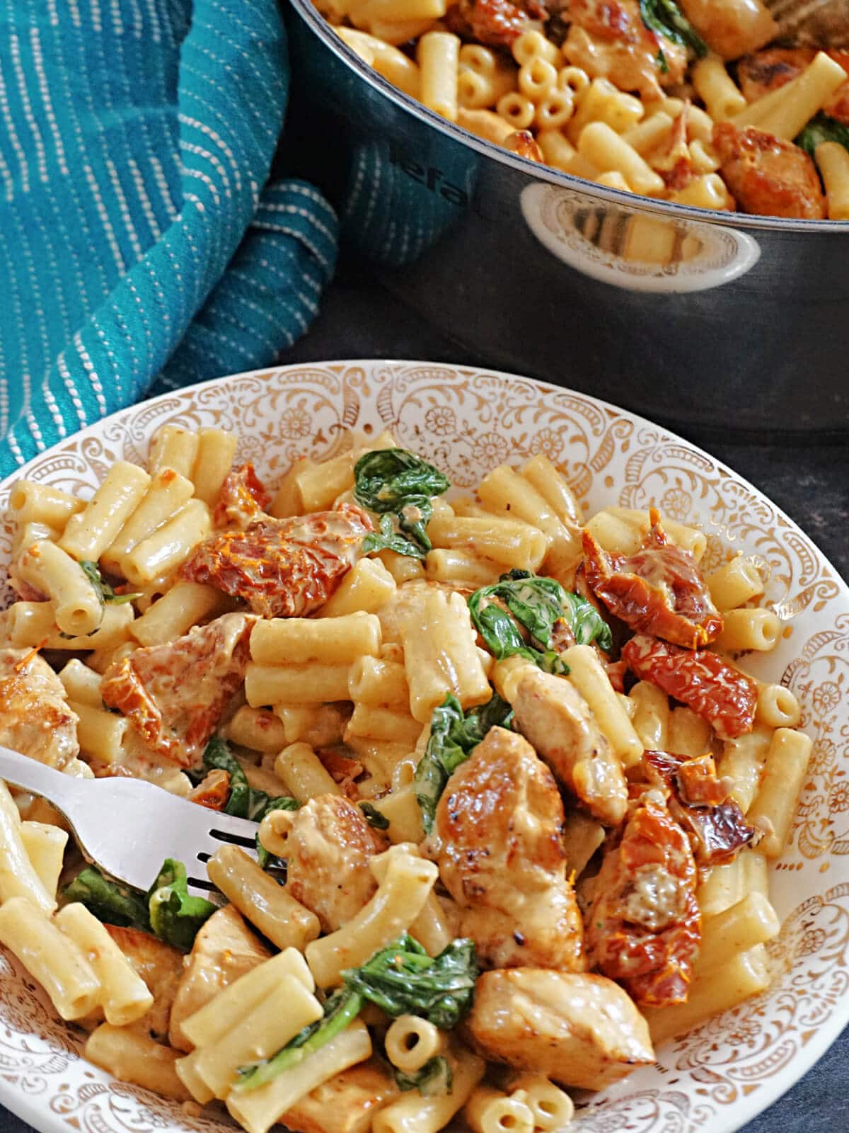 A plate with pasta with chicken, spinach and sun-dried tomatoes and a fork digging into it.