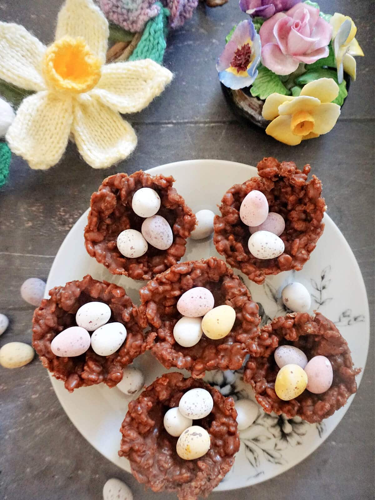 Overhead shot of a white plate with 6 chocolate rice krispie nests filled with mini chocolate eggs and decorations on the side