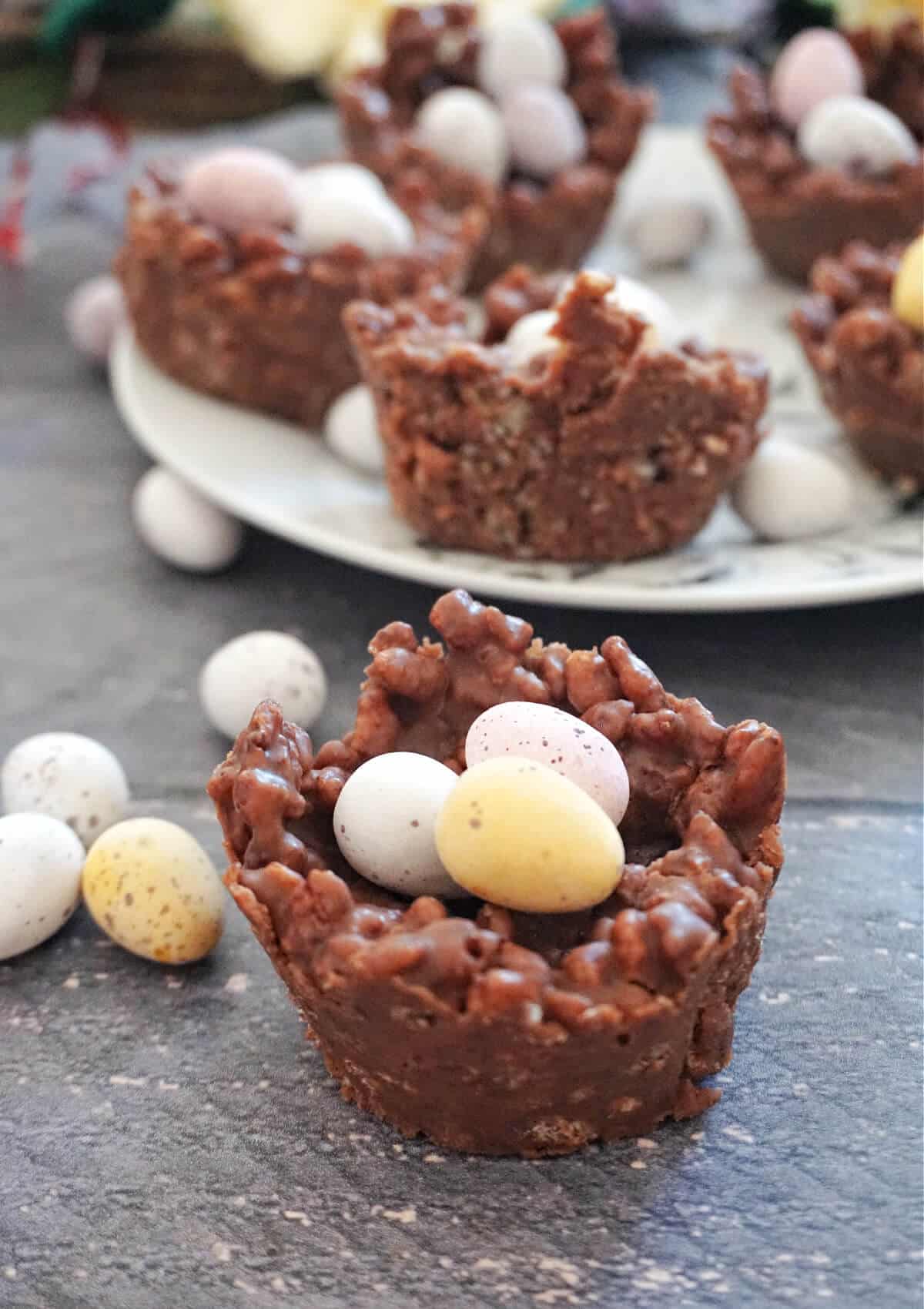 Close-up shot of a chocolate rice krispie nest filled with 3 mini chocolate eggs and a white plate with more nests in the background.