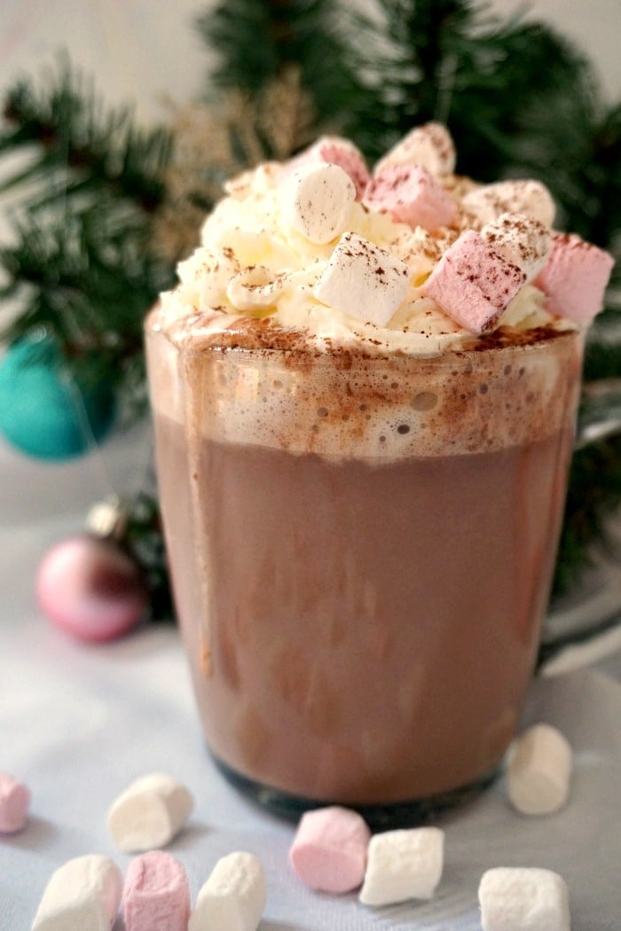 A glass of hot chocolate with marshmallows and cream with a Christmas tree in the background.