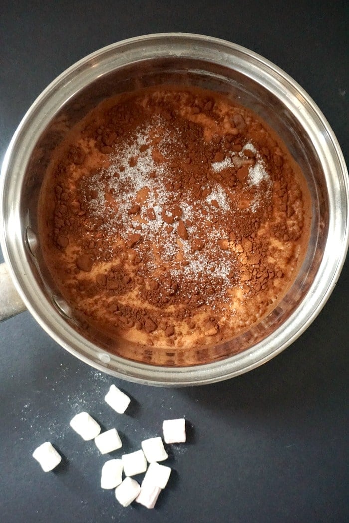 A pan of milk, cocoa powder and sugar on a black background with marshmallows next to it.