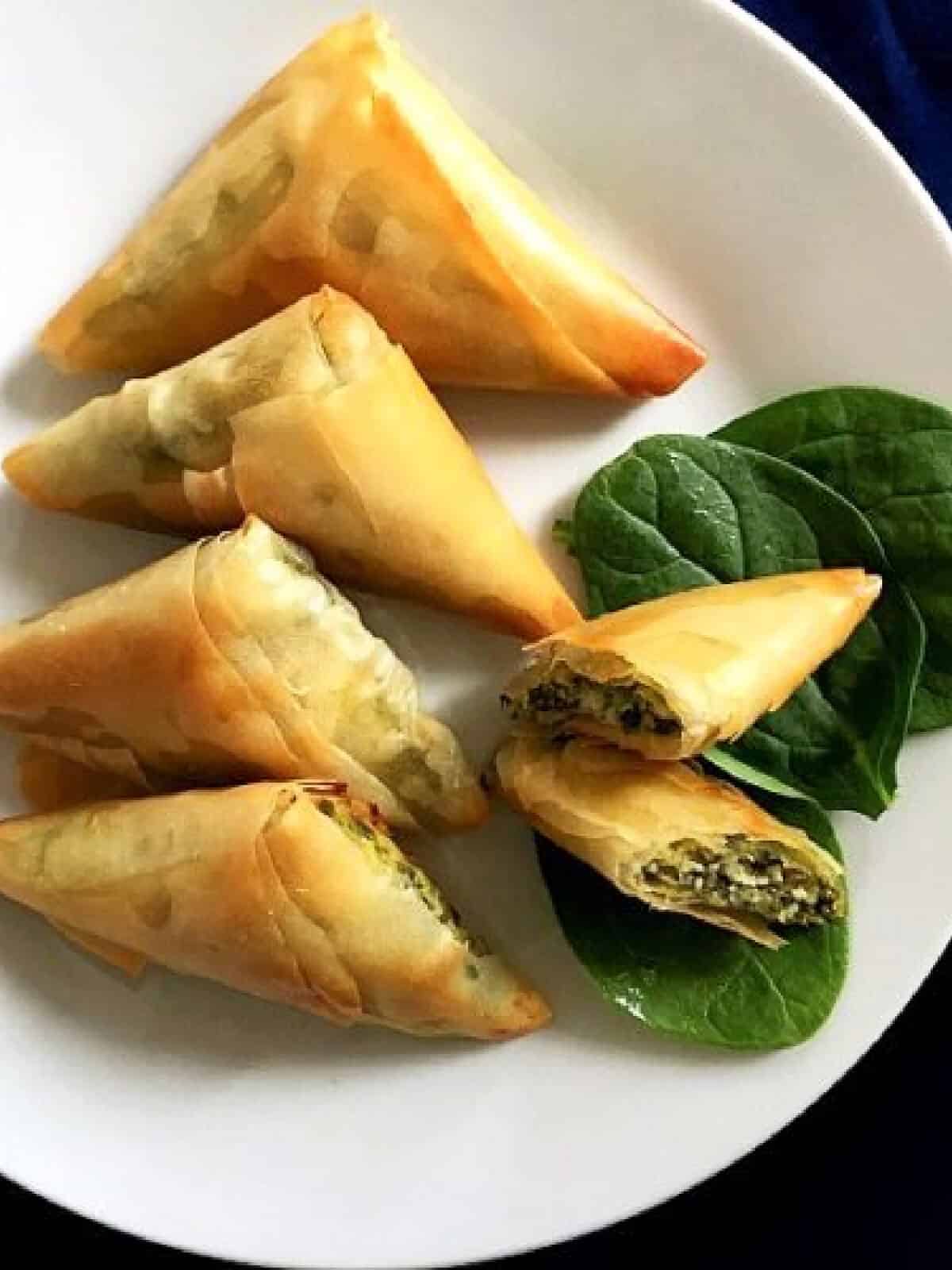 6 spanakopita triangles on a white plate with 3 spinach leaves.