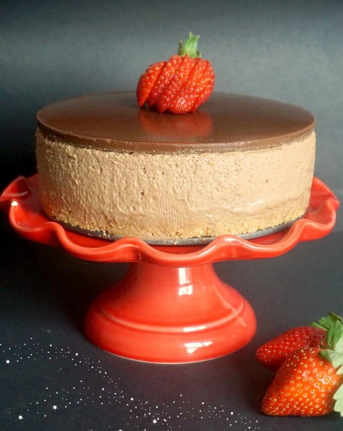 A red cake stand with a nutella cheesecake topped with a sliced strawberry and 2 strawberries next to the stand