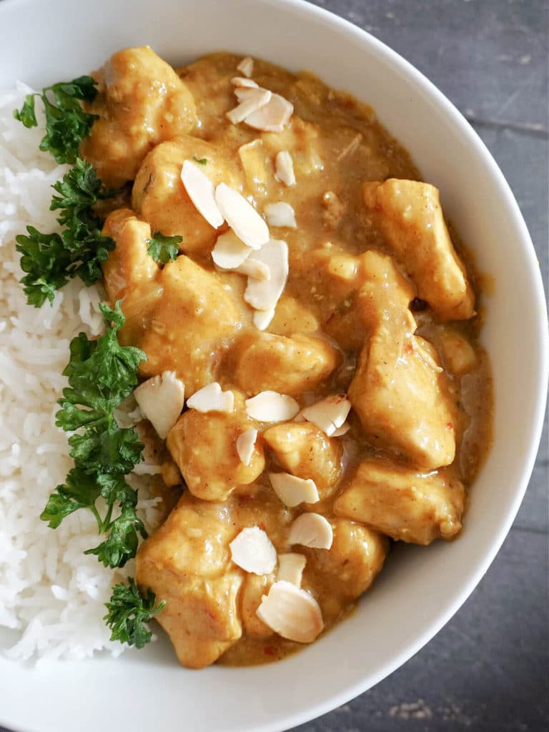 Close-up shot of a white plate with chicken curry topped with almonds and rice with parsley next to it