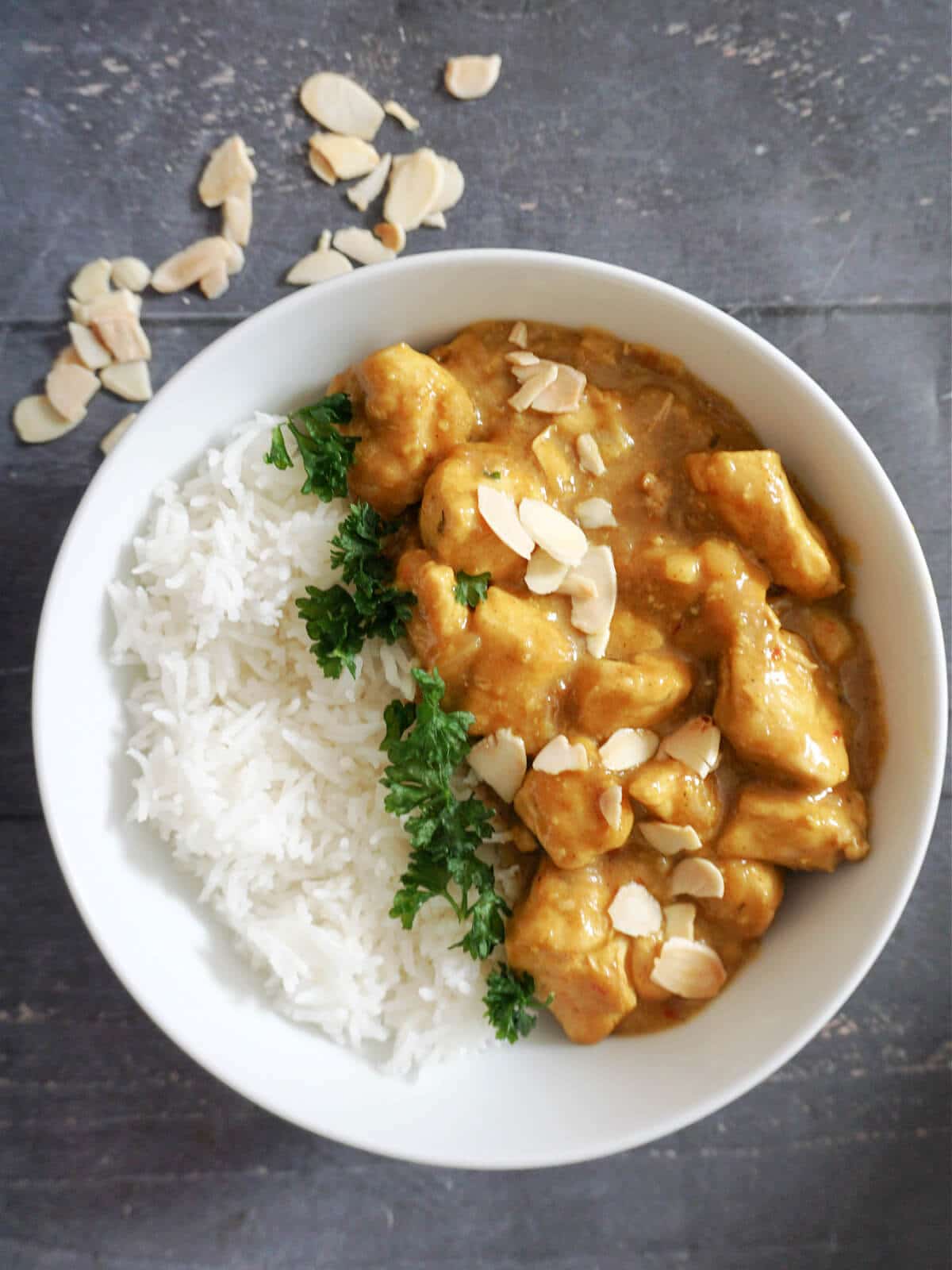 Overhead shoot of a white bowl with rice, chicken curry and parsley, with almonds scattered around