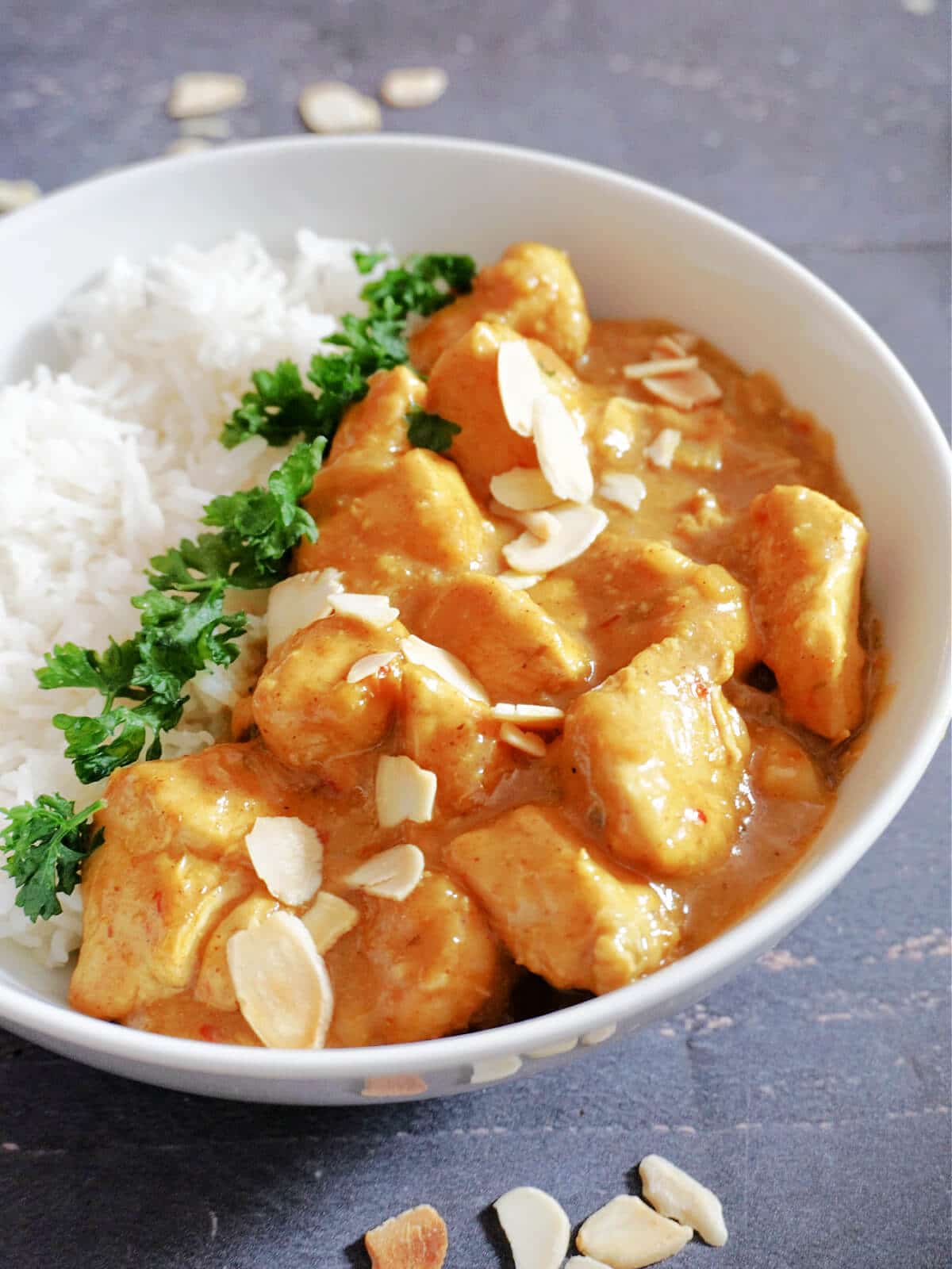 A white bowl with rice and chicken korma, garnished with parsley and almonds.