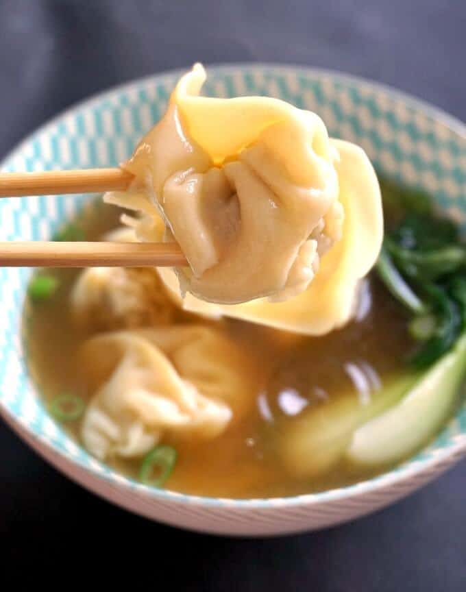 A wonton dumpling picked by chocpsticks, with a bowl of wonton soup in the background