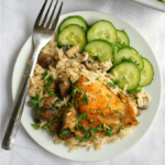 Overhead shoot of a white plate with baked chicken with rice and mushrooms and a cucumber salad on the side. Another pan with baked chicken with rice and mushrooms in the background