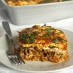 A white plate with a slice of high protein low carb zuccini lasagna and a fork on the side, and a lasagna dish in the background