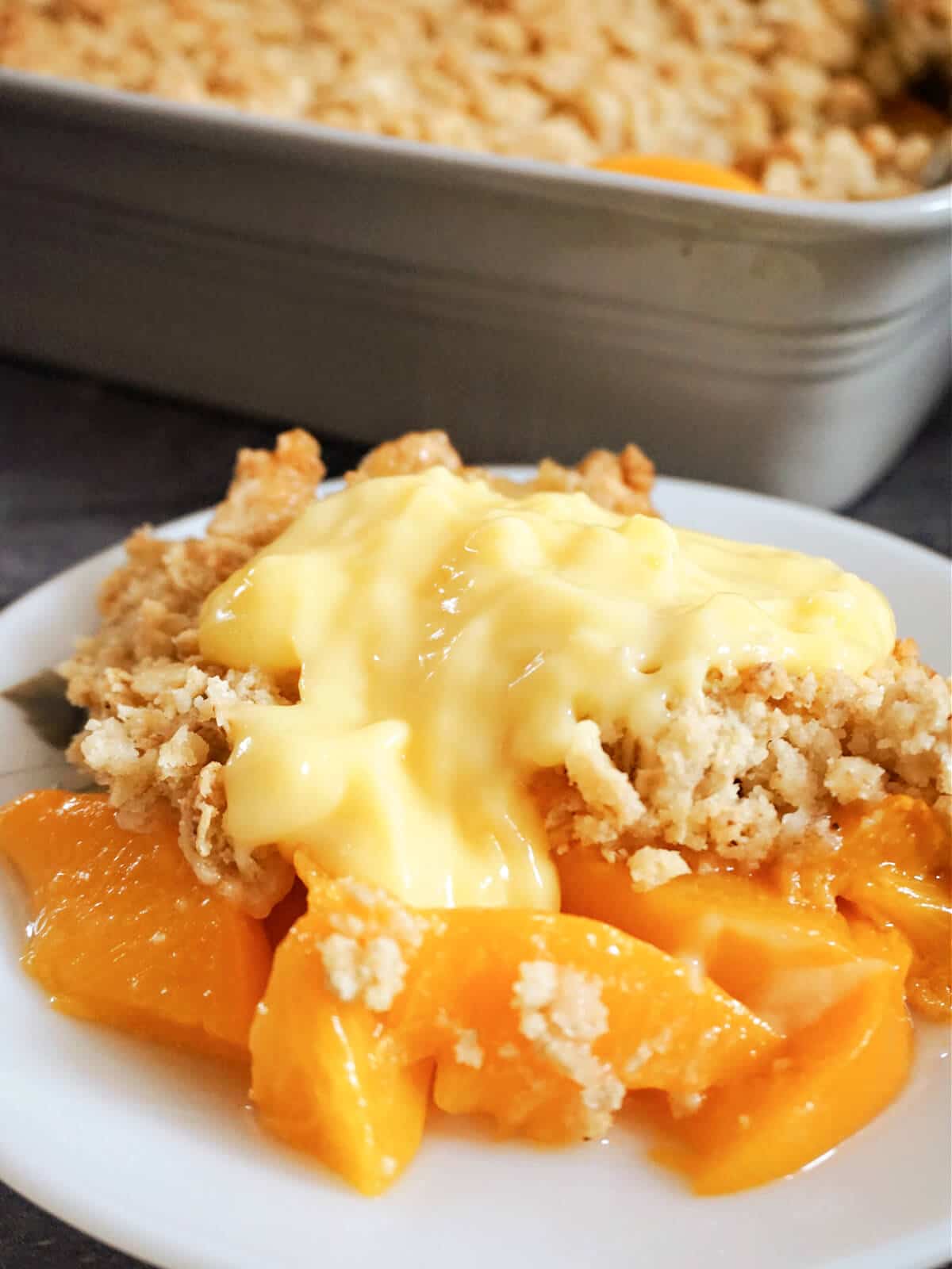 A white plate with peach crumble and a dish with more crumble in the background.