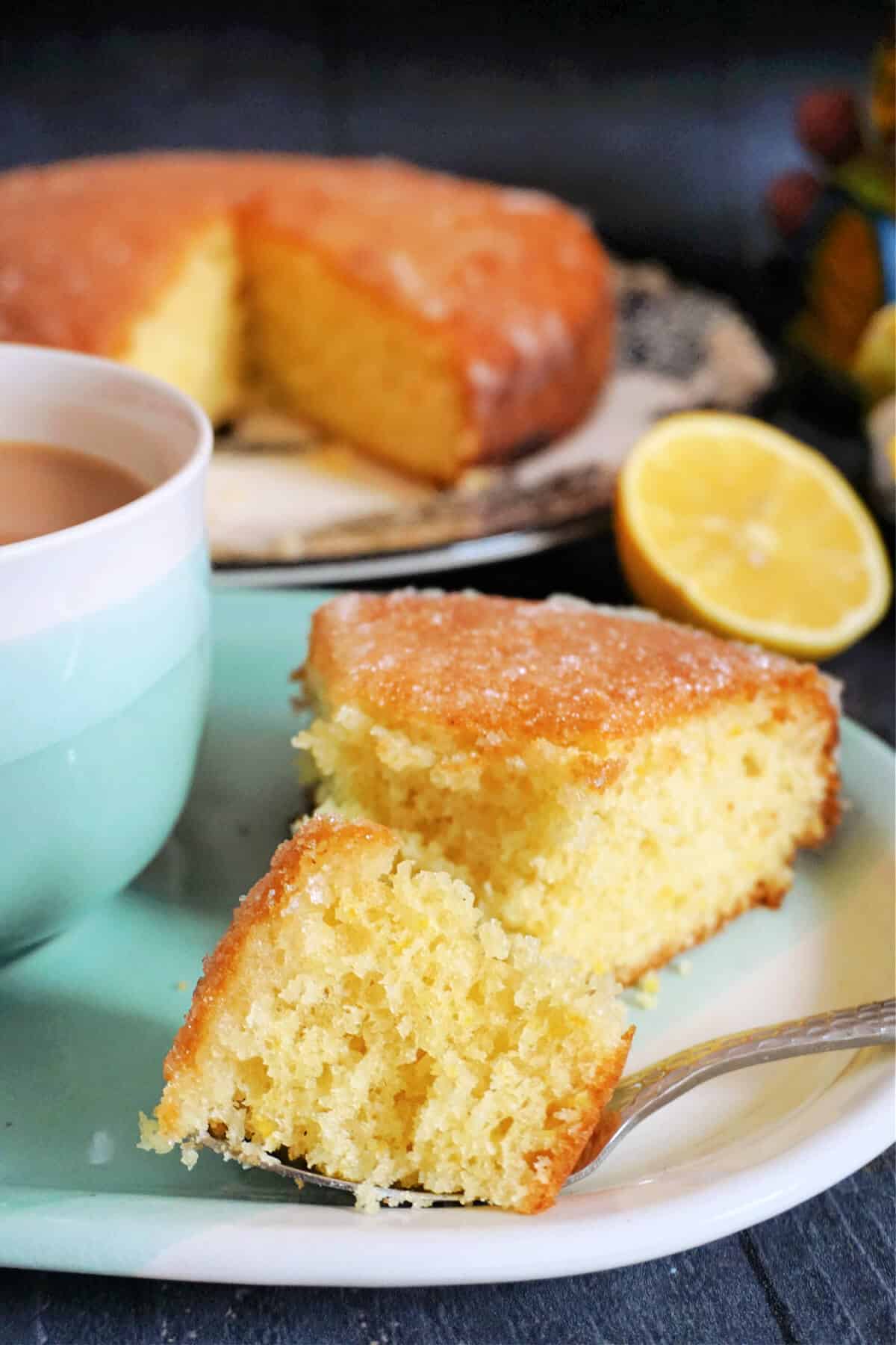 A slice of lemon drizzle cake on a light blue plate with half a lemon and more cake in the background