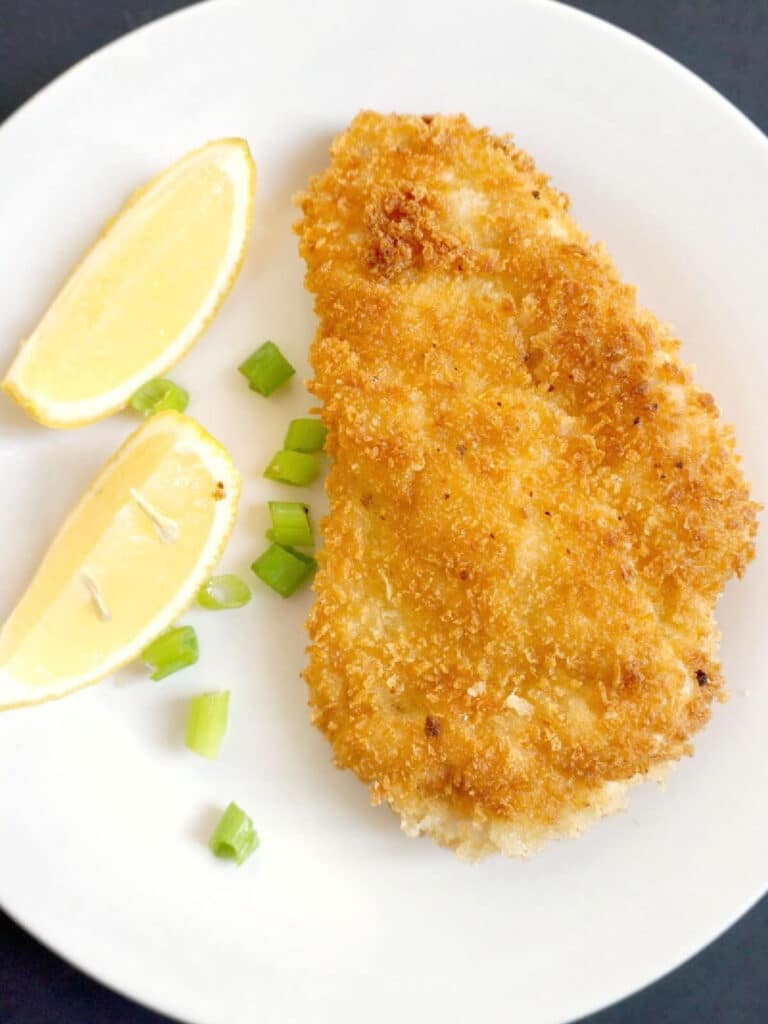 A white plate with a breaded chicken fillet, 2 lemon wedges and chopped spring onions