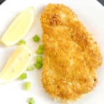 A white plate with a chicken schnitzel, 2 lemon wedges and chopped spring onions