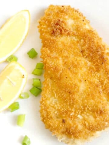 A white plate with a chicken schnitzel, 2 lemon wedges and chopped spring onions