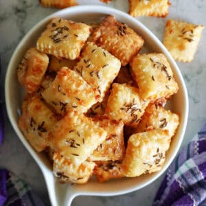 A white bowl with cheese crackers topped with salt and caraway seeds. More crackers outside the bowl.