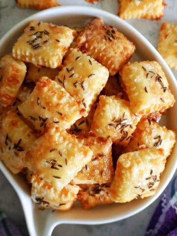 A white bowl with cheese crackers topped with salt and caraway seeds. More crackers outside the bowl.