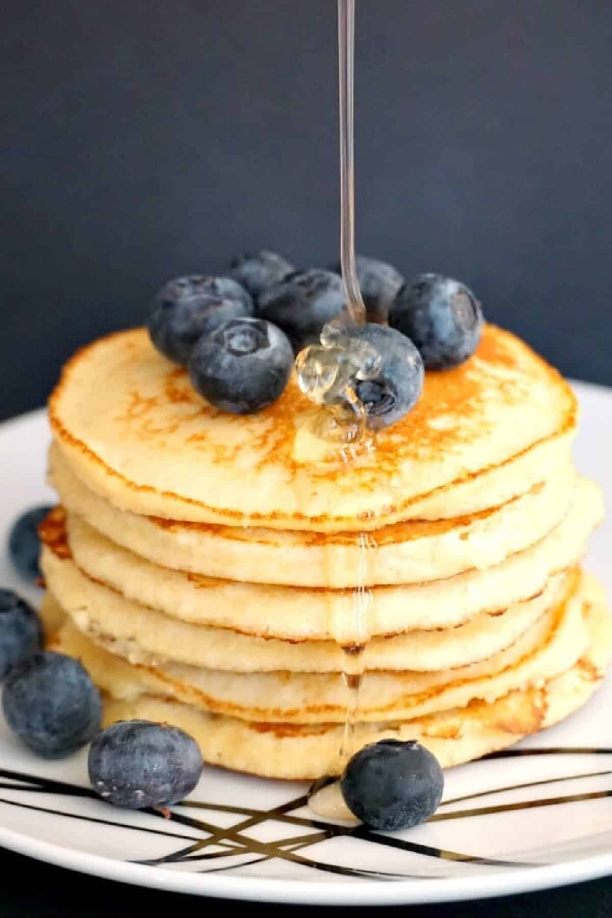 A pile of pancakes topped with blueberries and honey being drizzled over them.