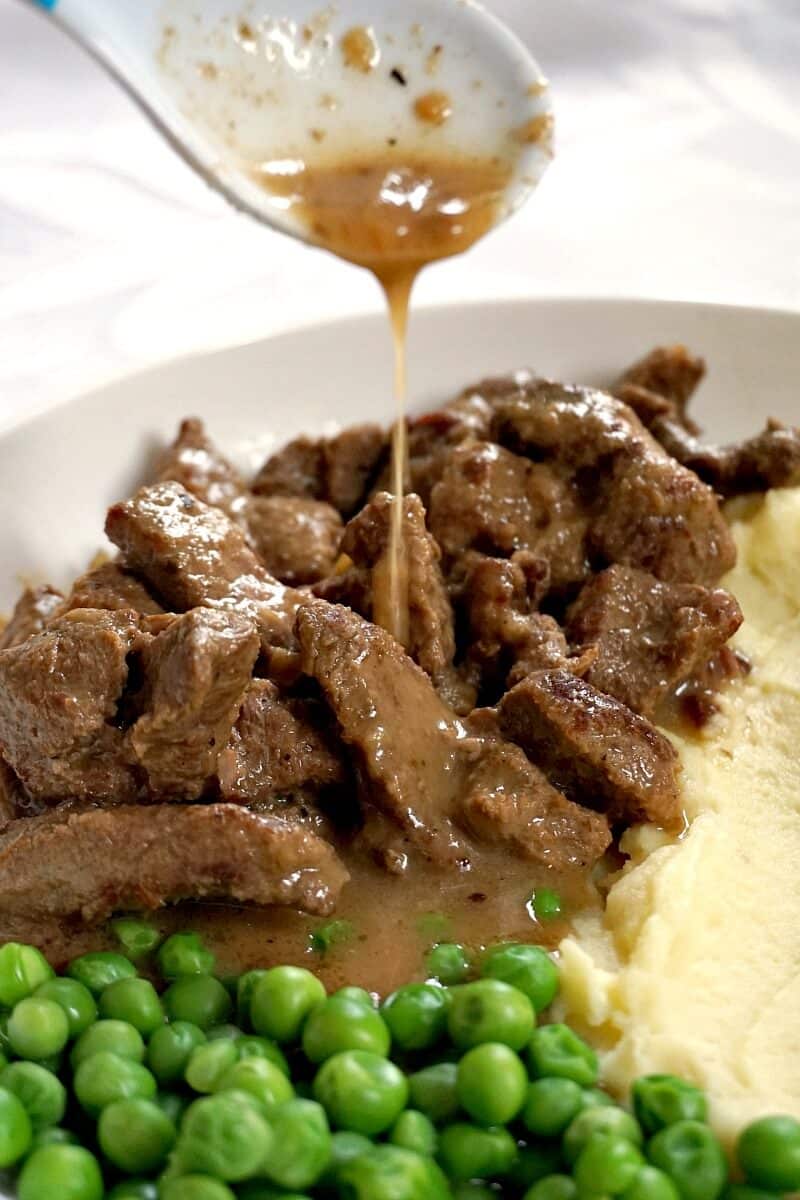 A white plate with beef tips, mashed potatoes and peas, and gravy being drizzled over the beef.