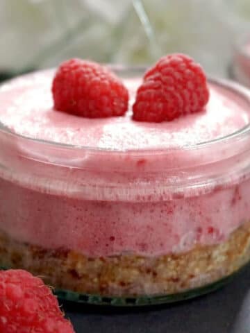 A glass ramekin with raspberry mousse cheesecake topped with 2 raspberries