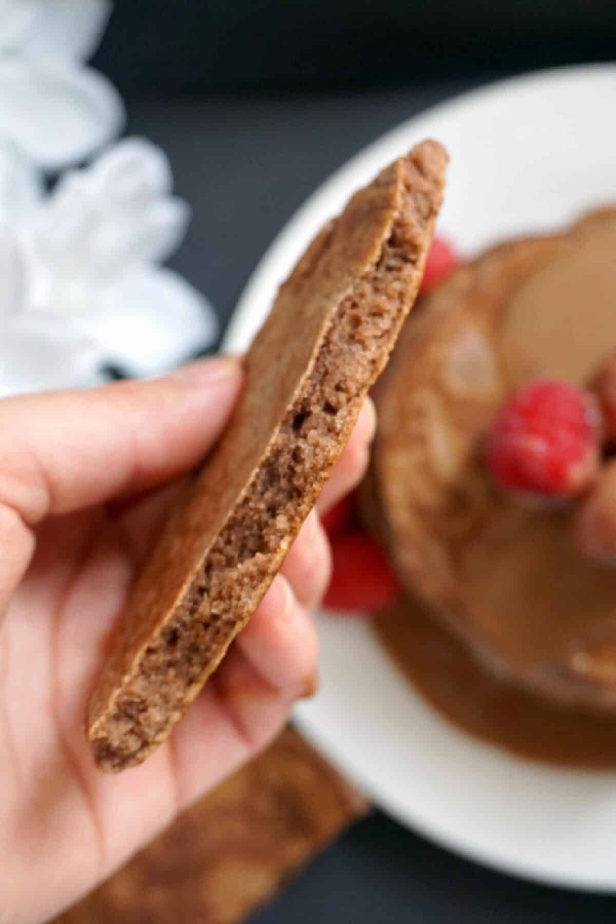 A half a chocolate pancake being held by hand with more pancakes in the background.