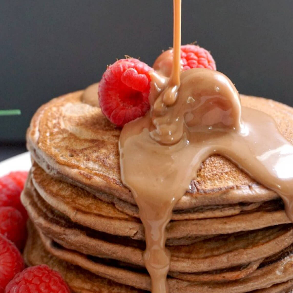 A pile of chocolate pancakes with 3 raspberries at the top and chocolate sauce being poured over them