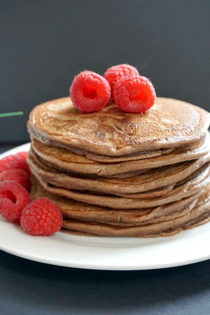 A stack of chocolate pancakes topped with 3 raspberries and more raspberries on the side of the plate