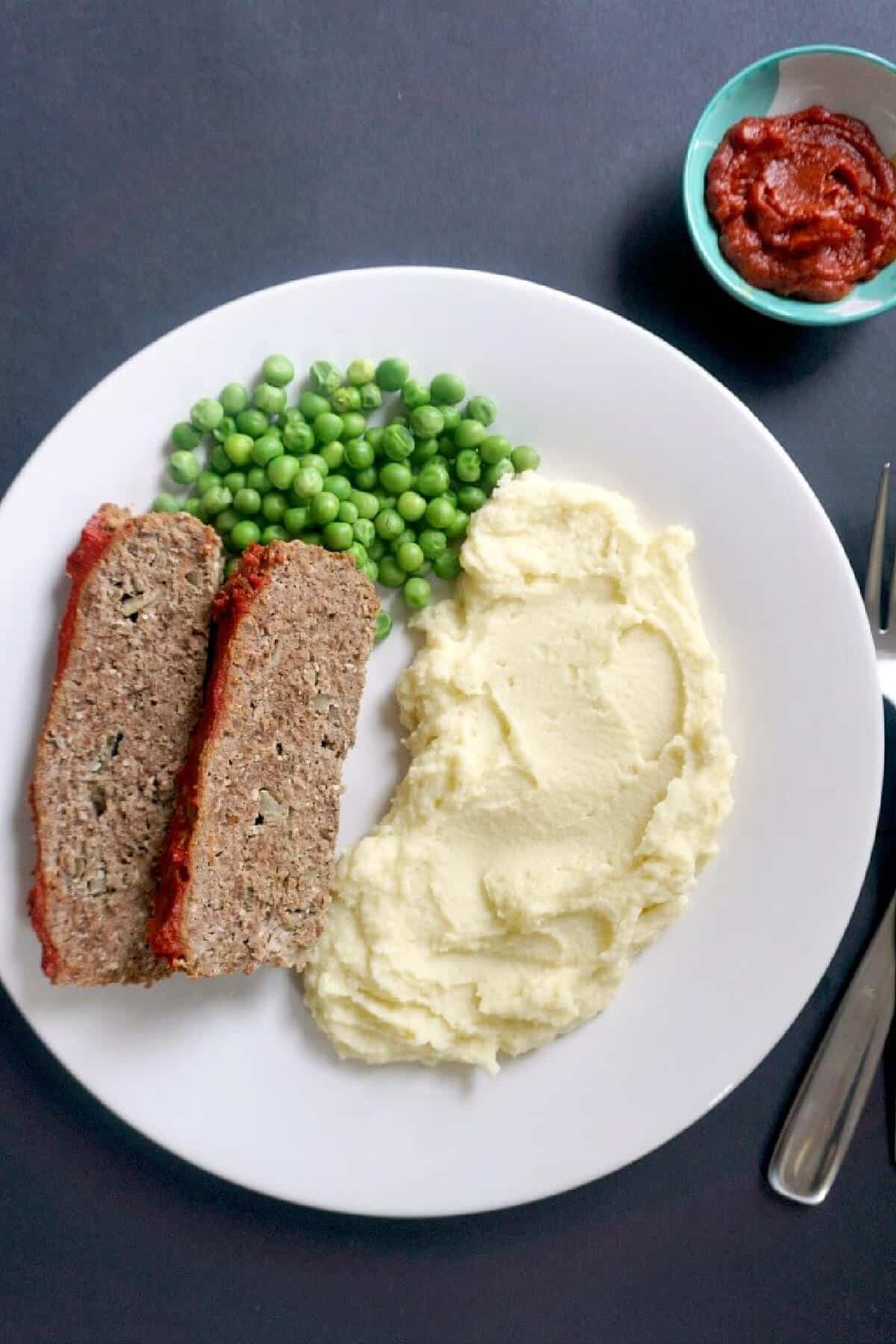 Overhead shoot of a white plate with 2 meatloaf slices, mash and peas.