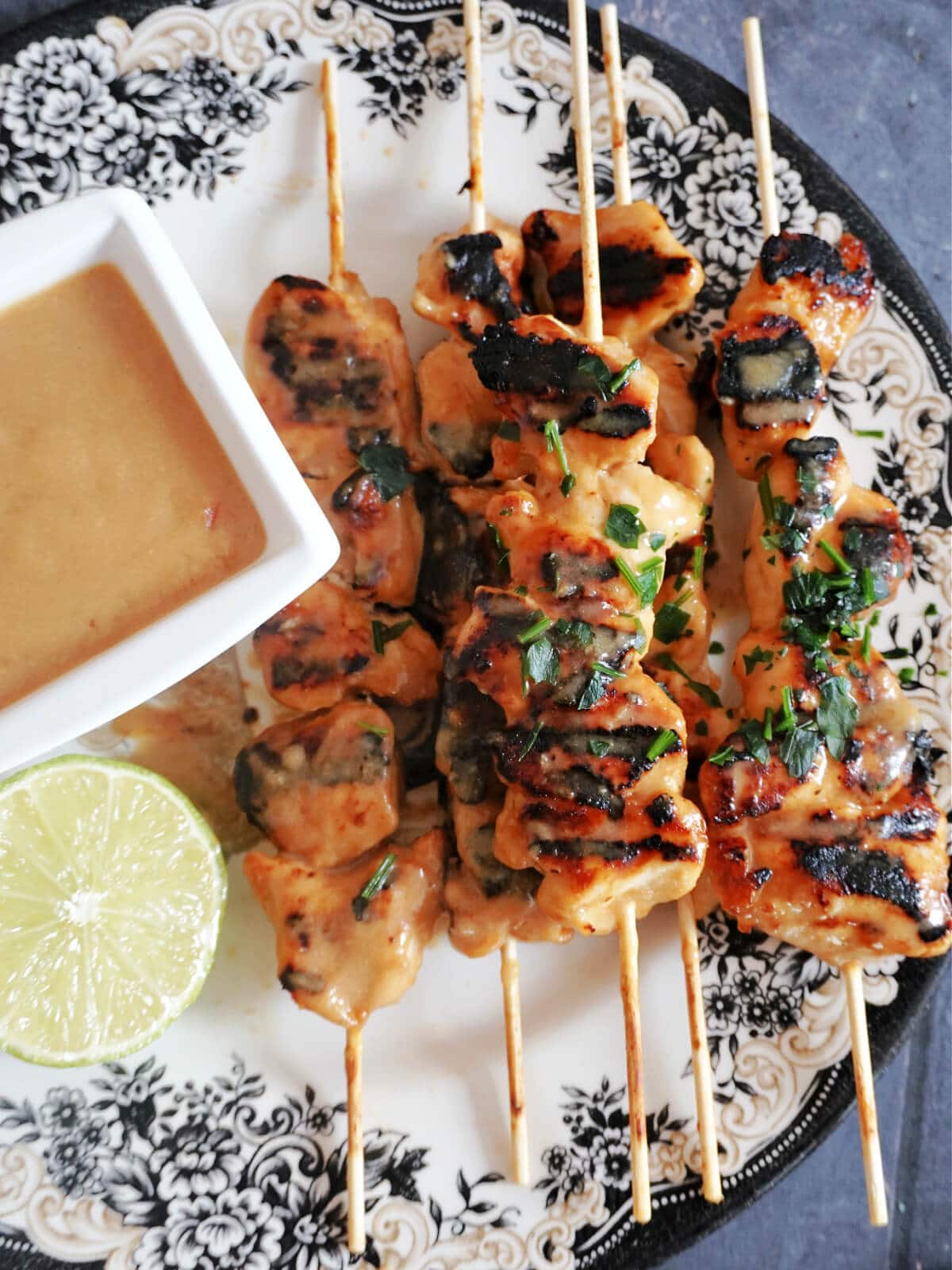 Overhead shot of grilled chicken skewers, a small bowl of peanut butter sauce, and half a lime on a white plate.