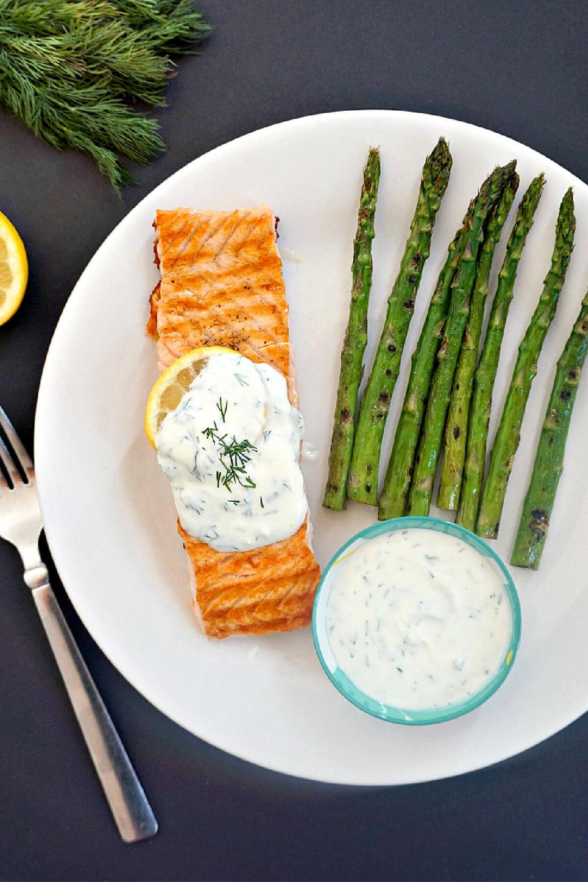 Overhead shoot of a white plate with a salmon fillet, asparagus and a ramekin with yogurt sauce