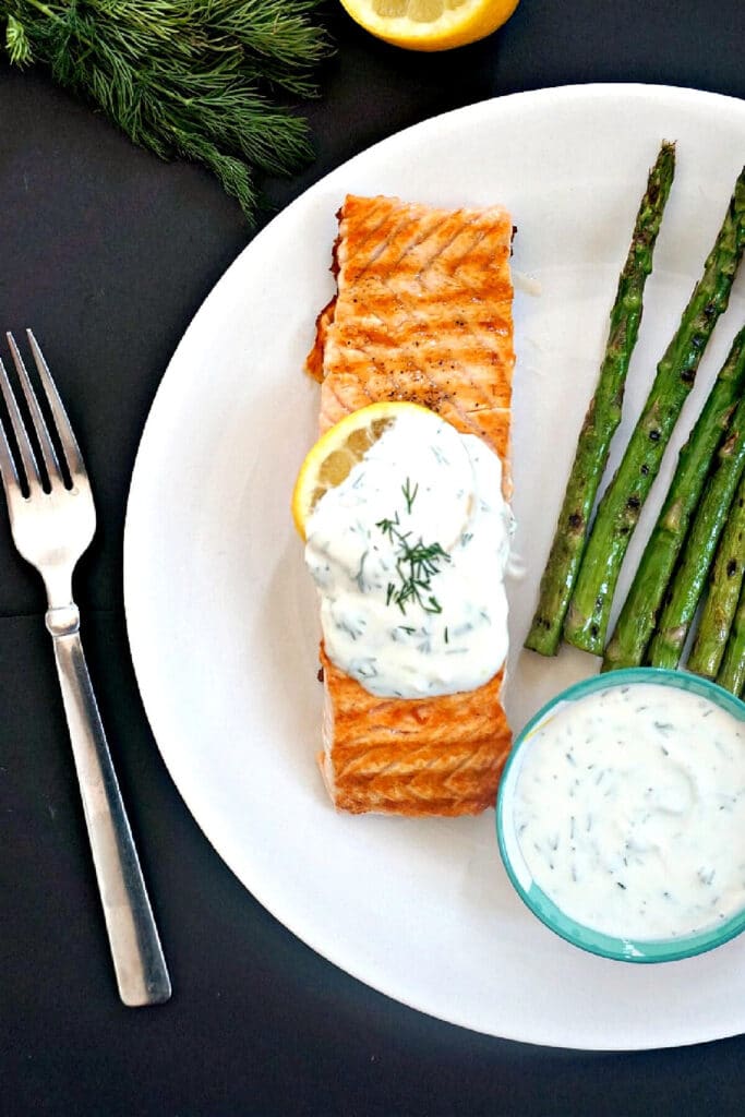 Overhead shoot of a white plate with a salmon fillet, grilled asparagus, a blue ramekin with yogurt sauce and a fork on the side