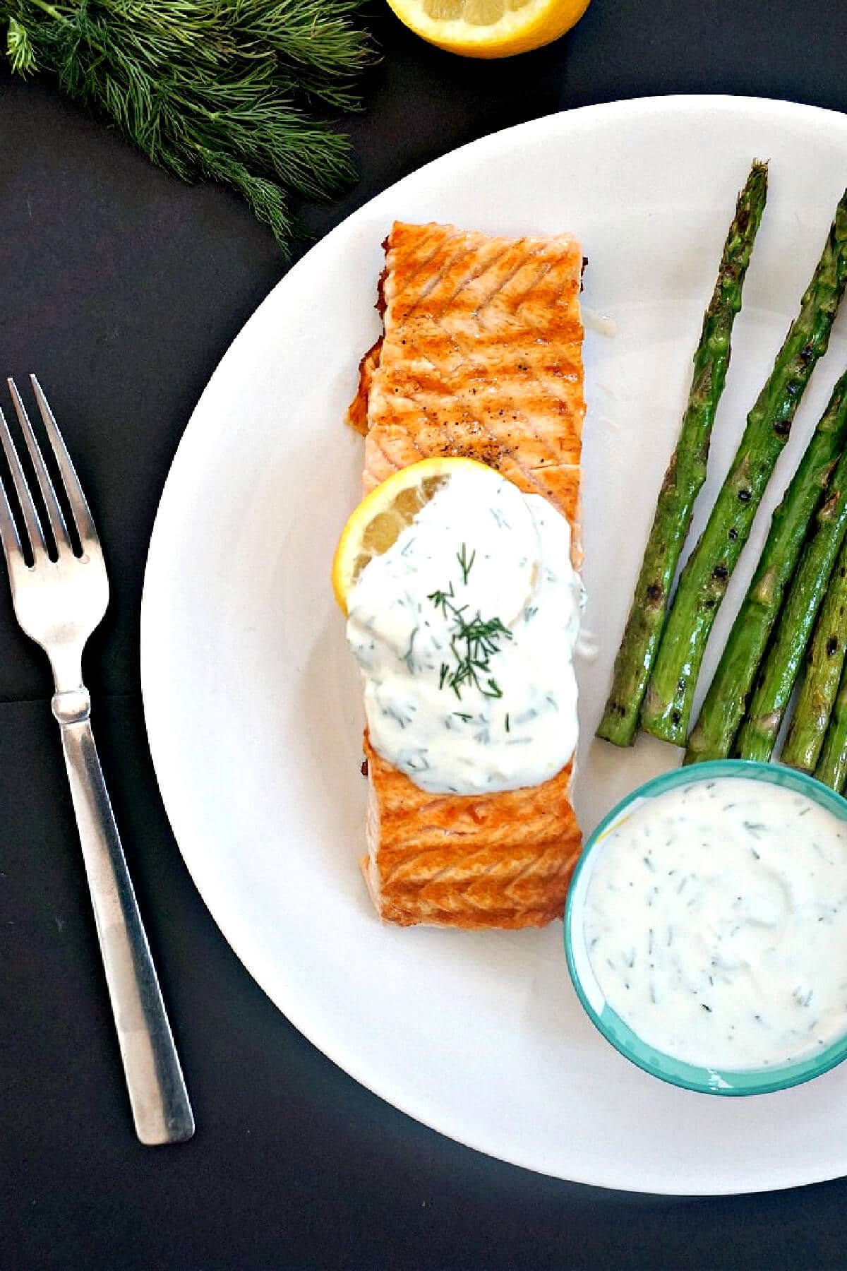 Overhead shoot of a white plate with a salmon fillet, grilled asparagus, a blue ramekin with yogurt sauce and a fork on the side.