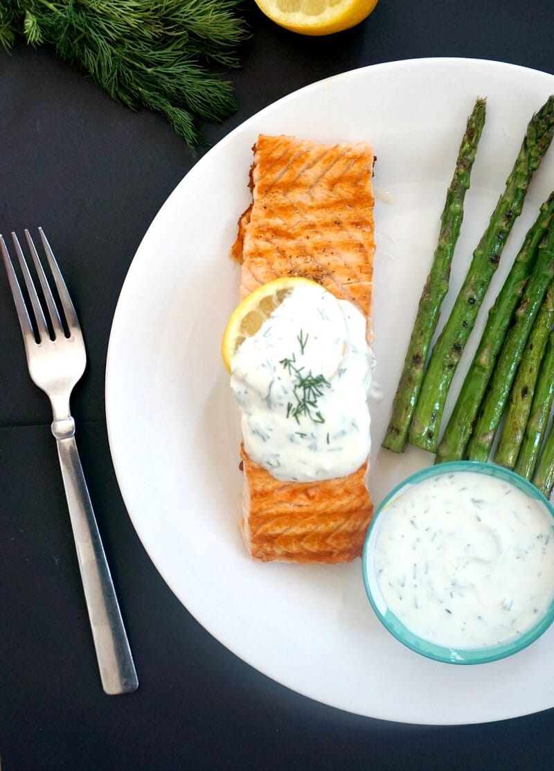 Half of a white plate with a grilled salmon fillet topped with yogurt dill sauce, grilled asparagus on the side, a fork and a bowl of sauce.