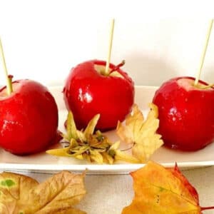 3 red candy apples on a white rectangle plate with yellow leaves around