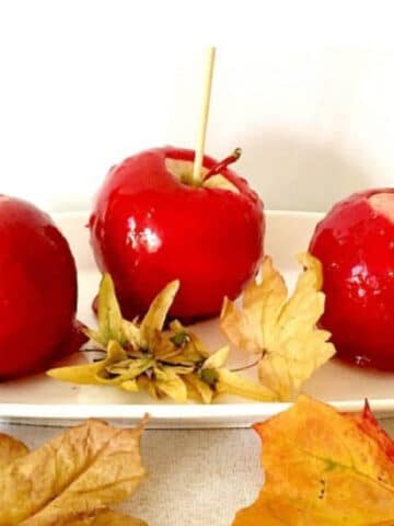 3 red candy apples on a white rectangle plate with yellow leaves around