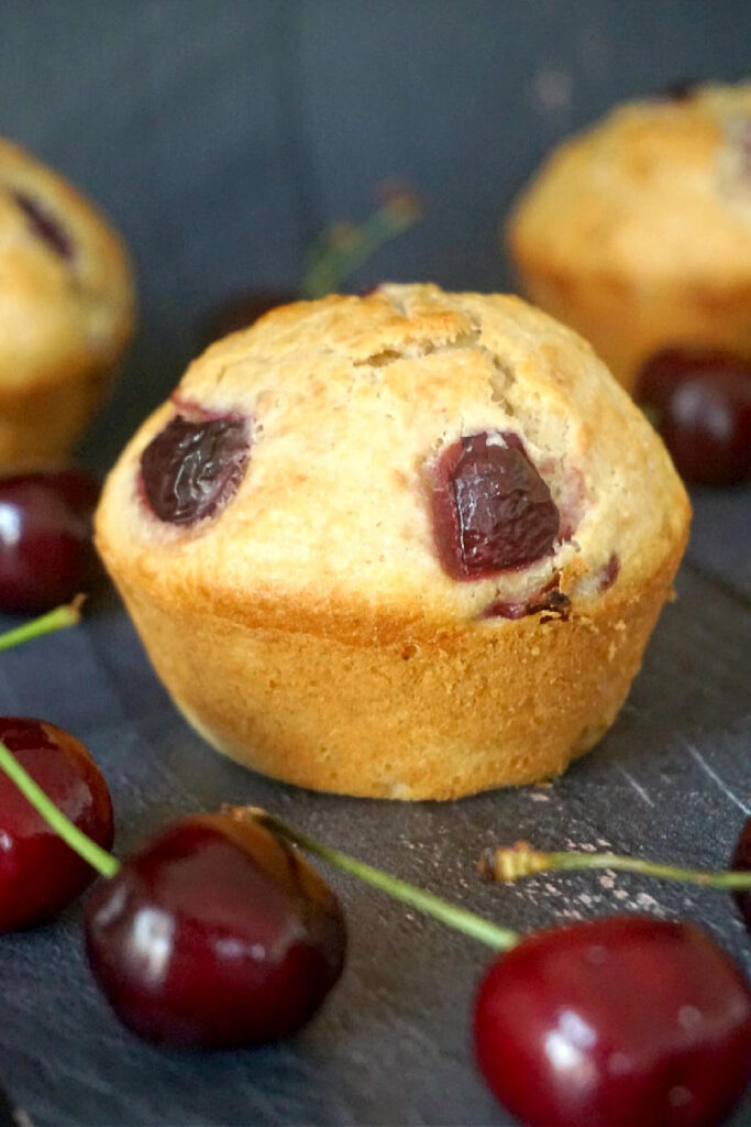 A cherry muffin with more muffins in the background and cherries around them