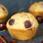 An almond cherry muffin with cherries around it and 2 other muffins in the background