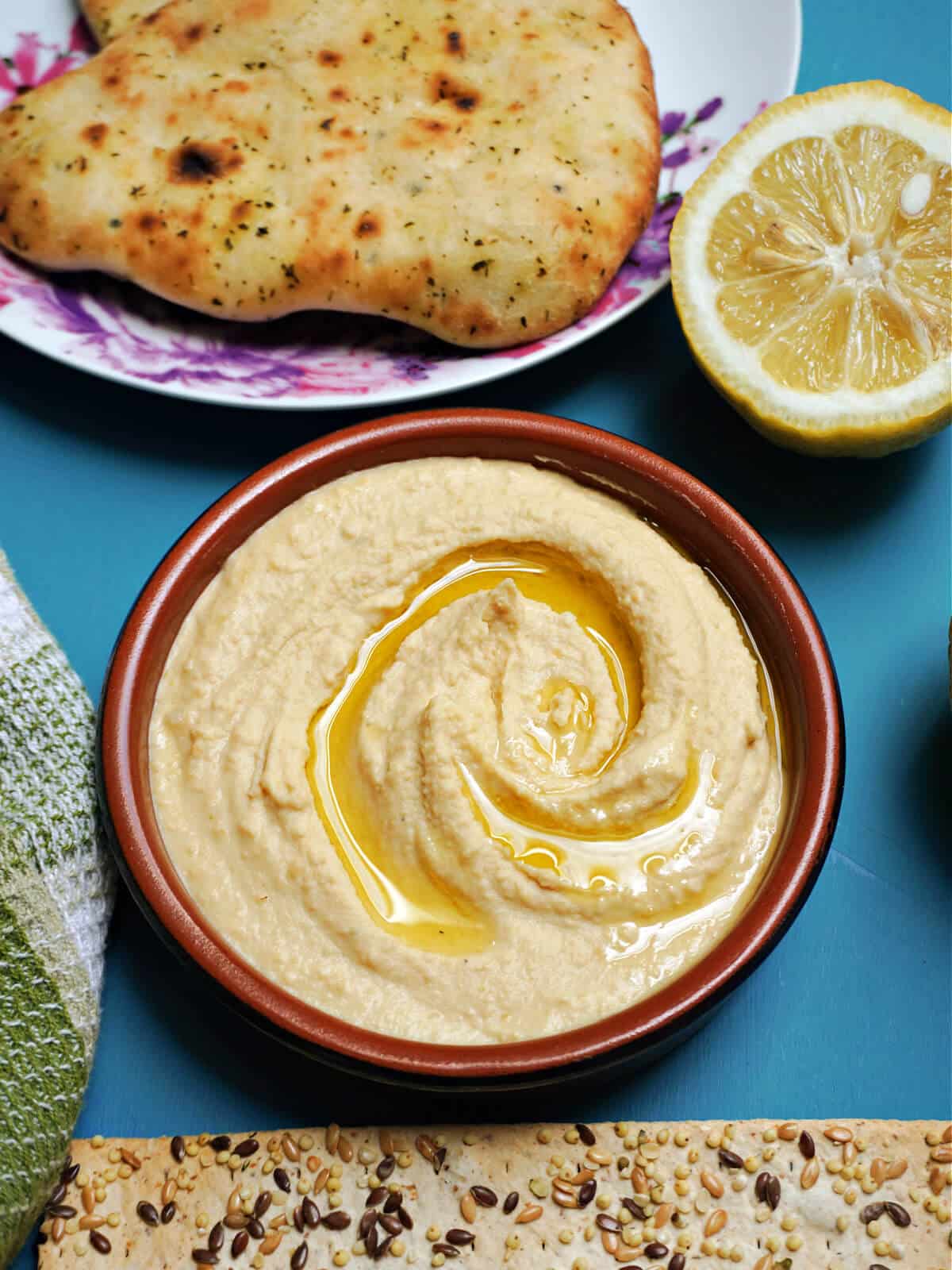 Overhead shoot of a brown ramekin with humus topped with olive oil, half a lemon and naan bread at the top.