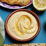 A brown ramekin with hummus drizzled with olive oil, half a lemon and naan bread at the top of the plate