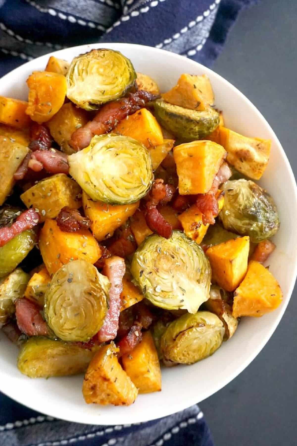 A white bowl with brussels sprouts, bacon and sweet potatoes.