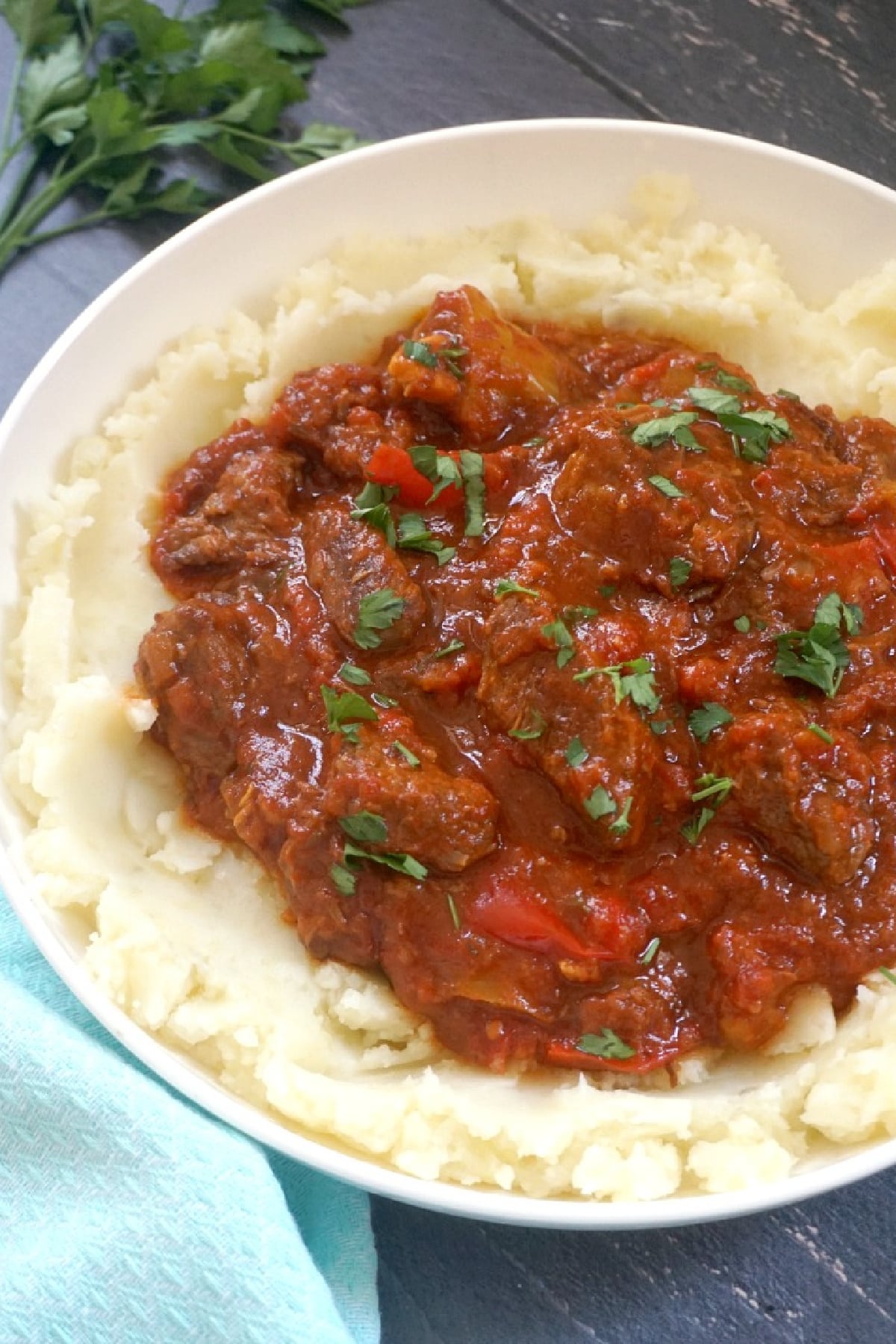 A white plate with beef goulash on a bed of mash.
