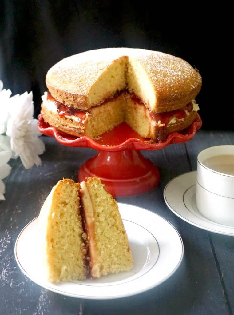 A slice of Victoria Sponge Cake on a white plate, a red cake stand with more cake and a cup of tea on the side