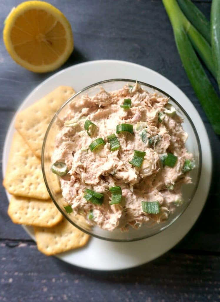 Overhead shoot of a glass bowl with tuna dip on a white plate with 4 crackers, half a lemon and spring onions on the side