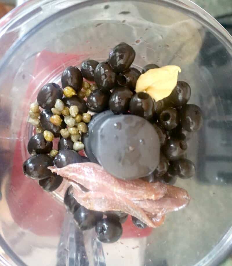 Overhead shoot of a blender with pitted black olives, anchovies filets, capers and dijon mustard