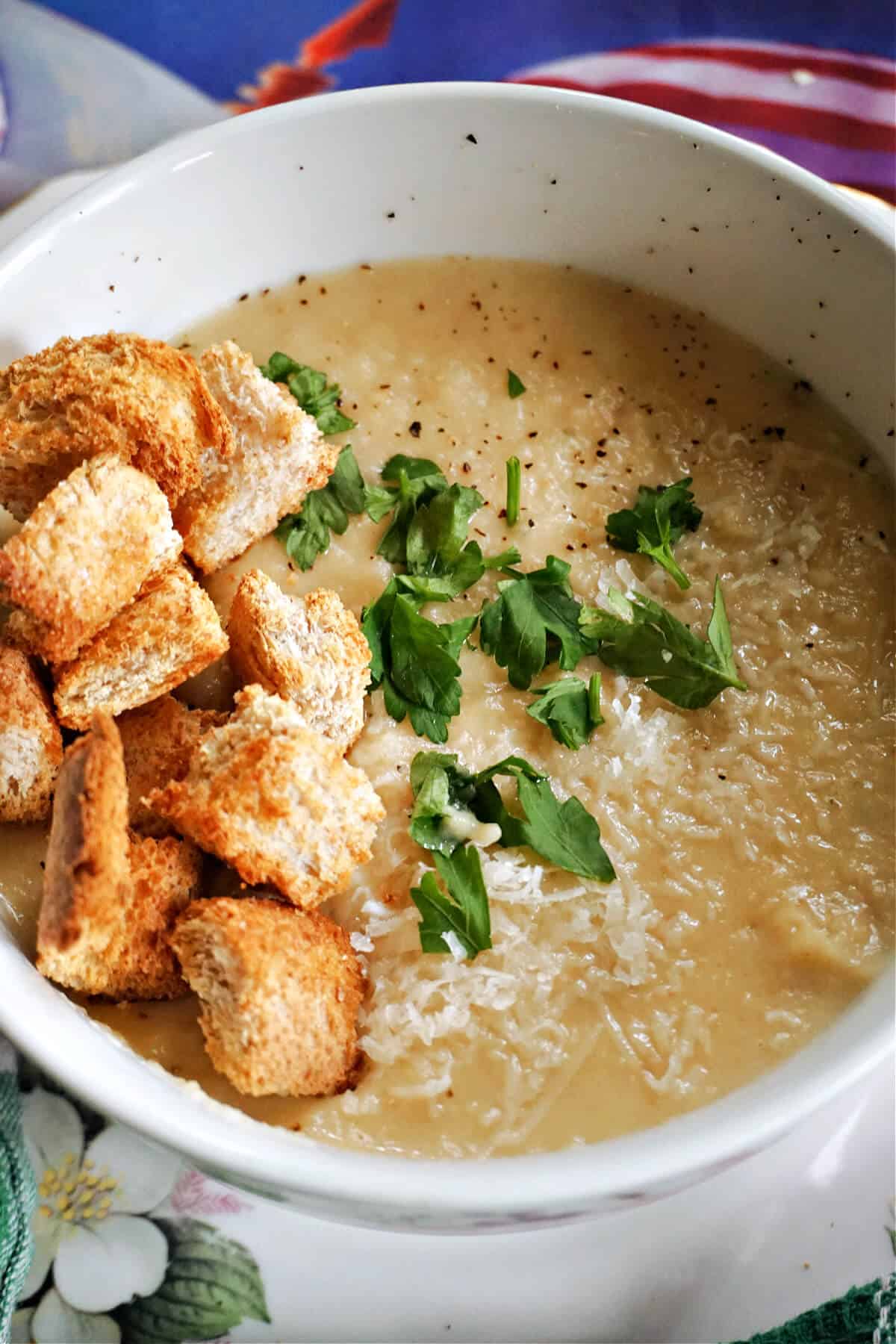 A white bowl with creamed soup topped with croutons, parmesan and chopped parsley.