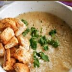 A white bowl with creamy parsnip soup topped with croutons, grated parmesan and chopped parsley.