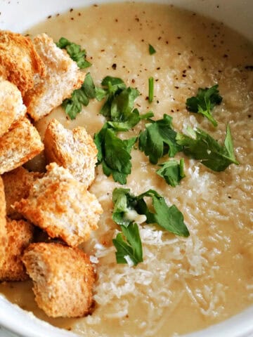 A white bowl with a portion of parnip soup topped with croutons, grated parmesan and chopped parsley.