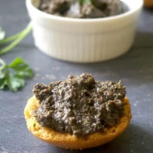 A slice of bread spread with tapenade and a white ramekin with more tapenade